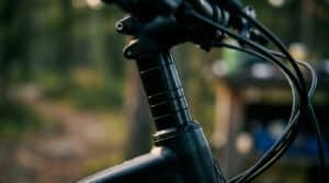 Détail d'un prolongateur de pivot de fourche de VTT noir, ajoutant de la hauteur. Bokeh extérieur vert/brun. Lumière naturelle. Suggère contrainte.